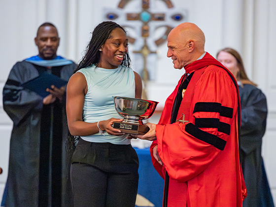 A smiling young woman with long braids, dressed in a light blue sleeveless top and dark pants, receives a large silver bowl trophy on a wooden base from a bald man wearing a bright red academic gown. Other individuals in black academic regalia stand in the blurred background.
