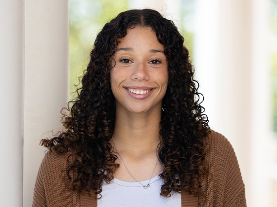 A smiling young woman with long, curly dark brown hair and freckles wears a white shirt, a brown cardigan, and a delicate silver necklace while posing for a headshot outdoors.