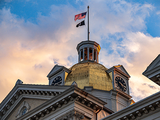 A low-angle view of a historic building's golden dome and clock tower, showcasing two visible clock faces under a dramatic evening sky lit with golden-hour clouds. An American flag and a black flag fly from a tall flagpole at the very top.