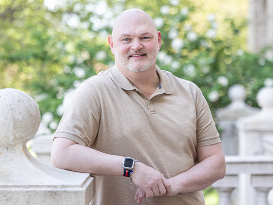 A portrait of a smiling, bald man with a light goatee, leaning against a stone balustrade outdoors while wearing a tan polo shirt and a smartwatch.