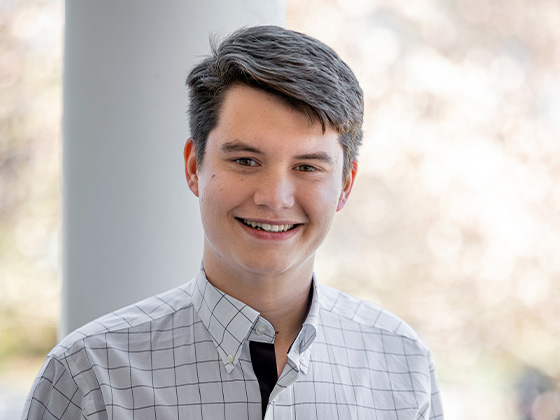 A smiling young man with short, neat dark hair poses for a headshot outdoors, wearing a white button-down shirt with a gray grid pattern.