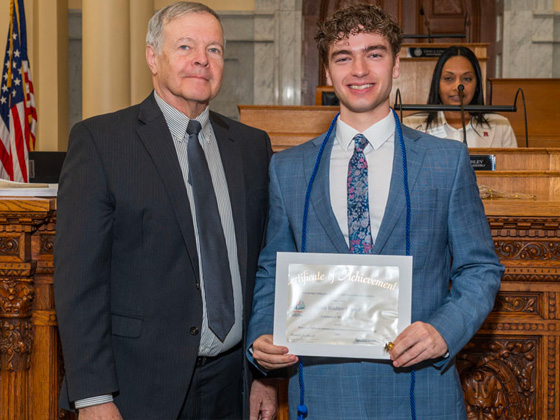 A young man with curly brown hair in a blue patterned suit and floral tie smiles while holding a "Certificate of Achievement" next to an older man in a dark suit and tie. They are standing indoors in a formal hall with an American flag, wooden desks, and an ornate wooden bench in the background.