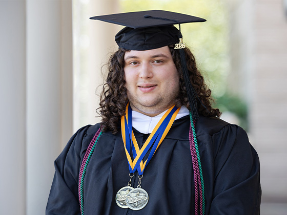 A portrait of a male graduate with long curly brown hair, wearing a black cap and gown, academic honor cords, and two medals with yellow and blue ribbons around his neck.