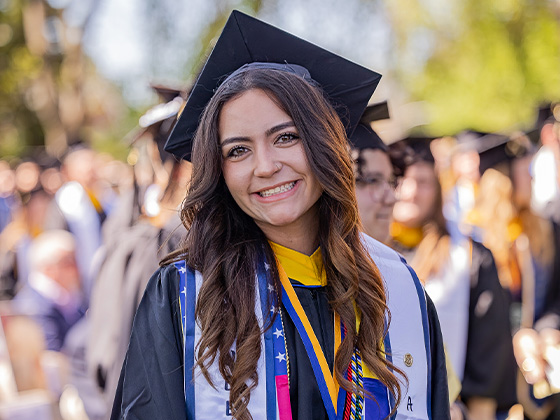 A close-up portrait of a smiling female graduate with long brown hair, wearing a black cap and gown, academic stoles, and medals at an outdoor commencement ceremony.