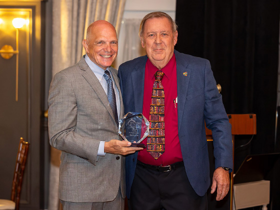 Two men standing indoors, smiling at the camera as the man on the left presents a crystal award to the man on the right.
