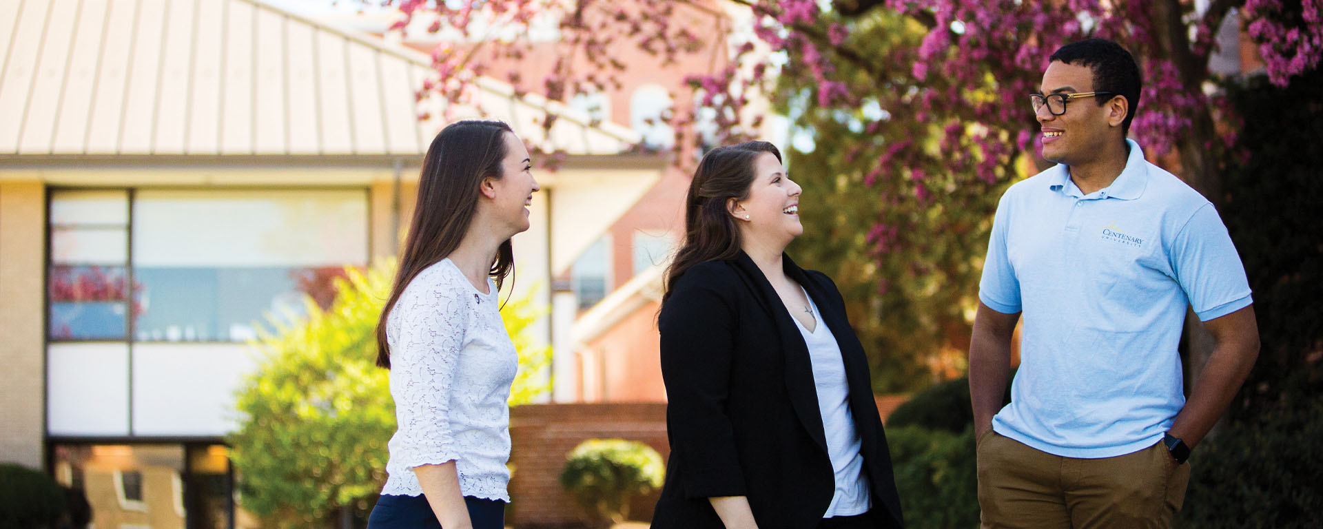 lifeatcent Three smiling university students standing outside on campus chatting, with a building and flowering pink trees in the background.