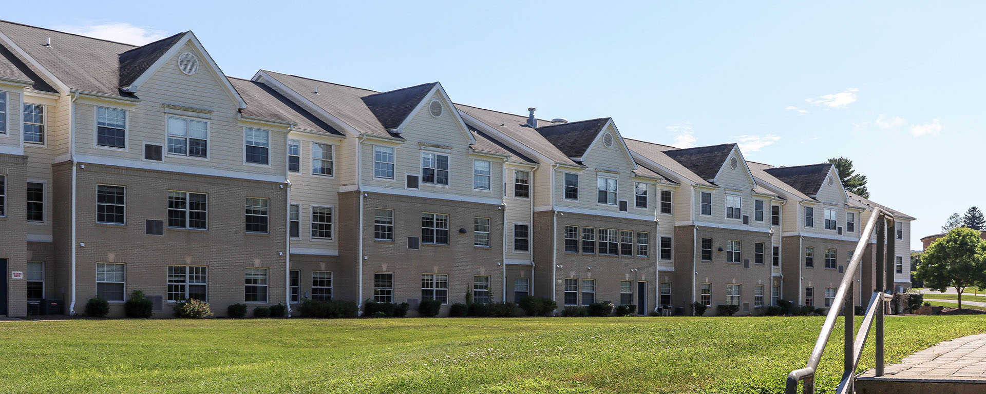 A long, modern multi-story student residence hall building with beige siding and brick accents, facing a wide green lawn on a clear day.