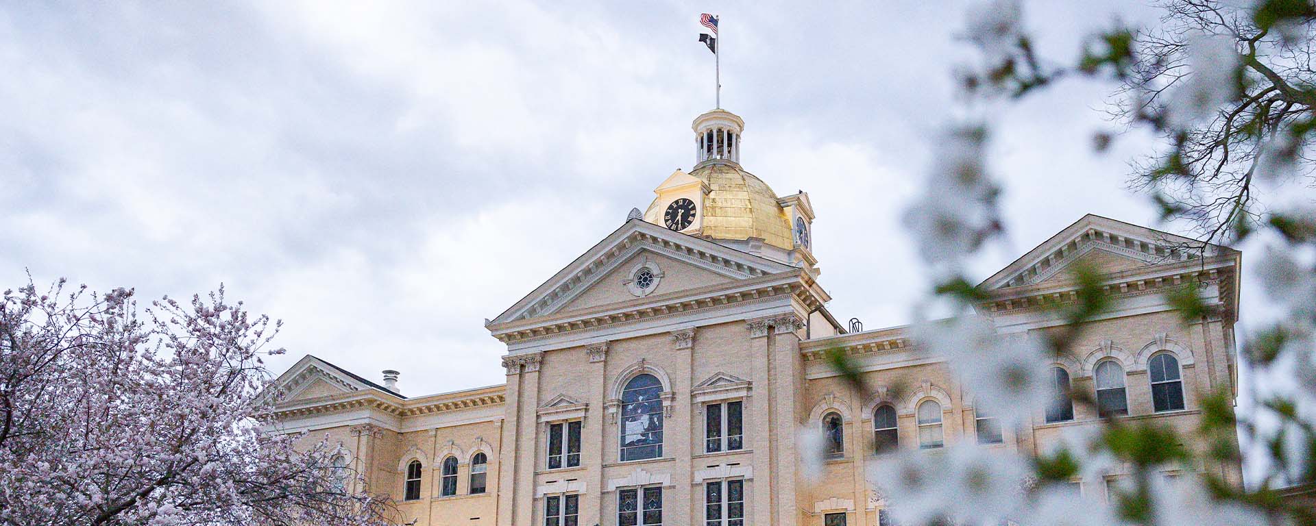 ssvp The historic, brick Edward W. Seay Administration Building featuring its prominent gold dome and clock tower under a cloudy sky, partially framed by blooming spring tree blossoms in the foreground.