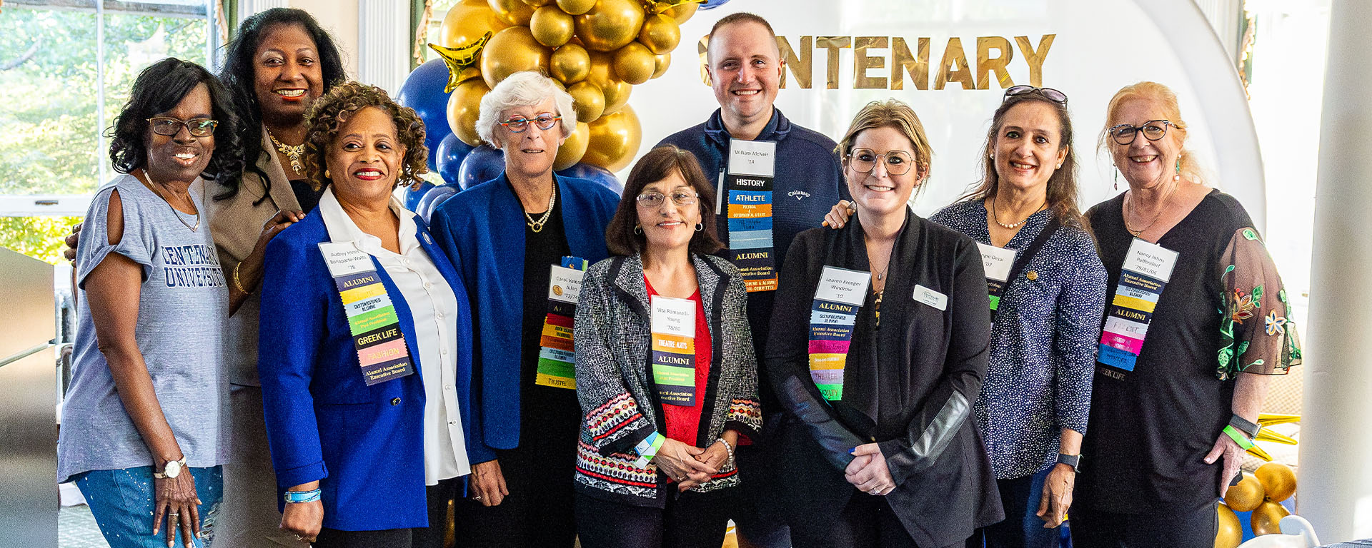 A group of eight smiling people standing together at a Centenary University event, with gold and blue balloons in the background.