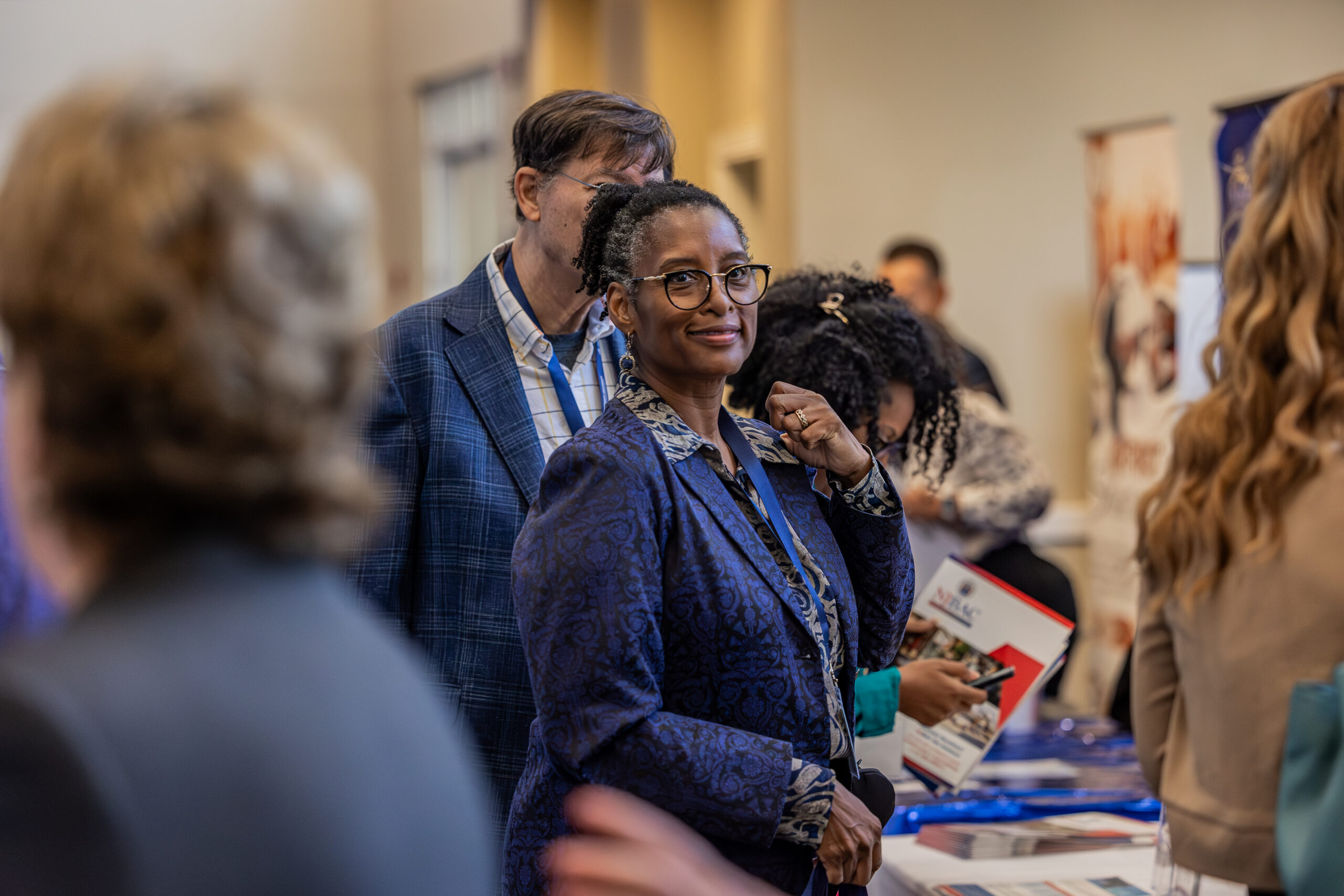 A woman looks at a camera while she stands at a vendor table surrounded by other people.
