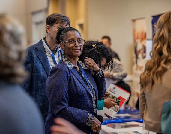 A woman looks at the camera while standing near a vendor table in a crowded room.