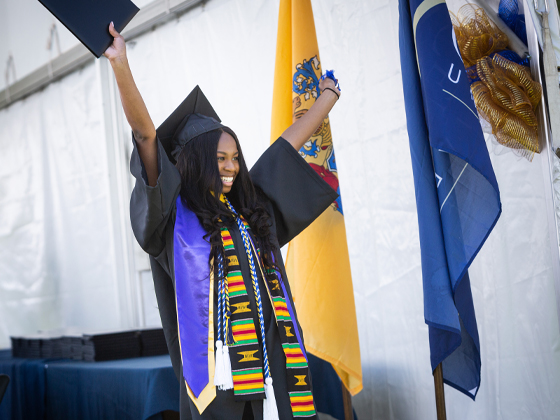 A woman in graduation regalia throws her hands up in the air, celebrating receiving her diploma.