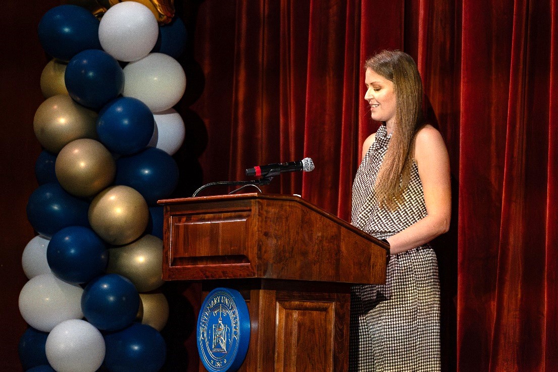 A woman stands at a podium in front of a red curtain and balloons.