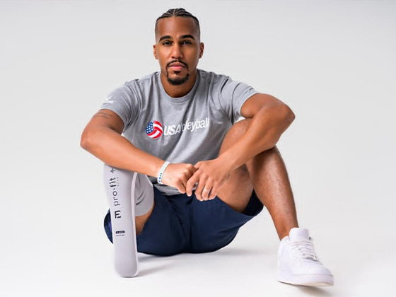 A Black man with one leg sits on the floor wearing a gray USA volleyball shirt.