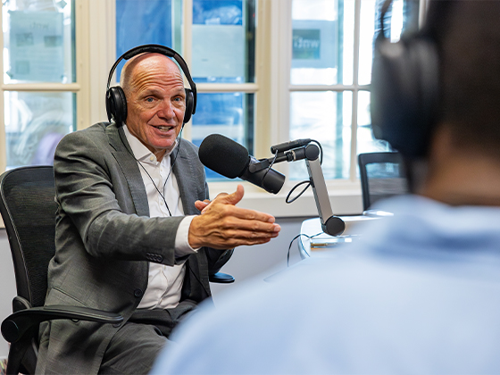A man in a suit wearing headphones speaks into a microphone and gestures to a person interviewing him.