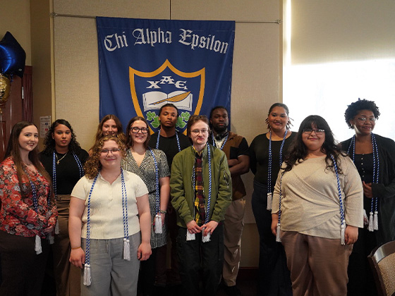 A group of ten diverse college students wearing blue and white honor cords stands in front of a large blue Chi Alpha Epsilon banner.